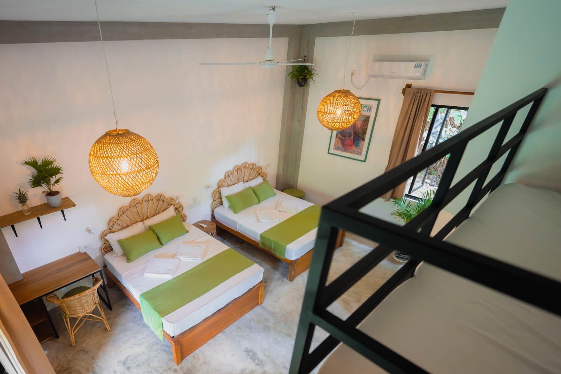 Ein Schlafzimmer mit zwei Betten, einem Schreibtisch und einem Stuhl. Zwei Pendelleuchten, ein Deckenventilator und ein Fenster mit Vorhängen sind vorhanden.