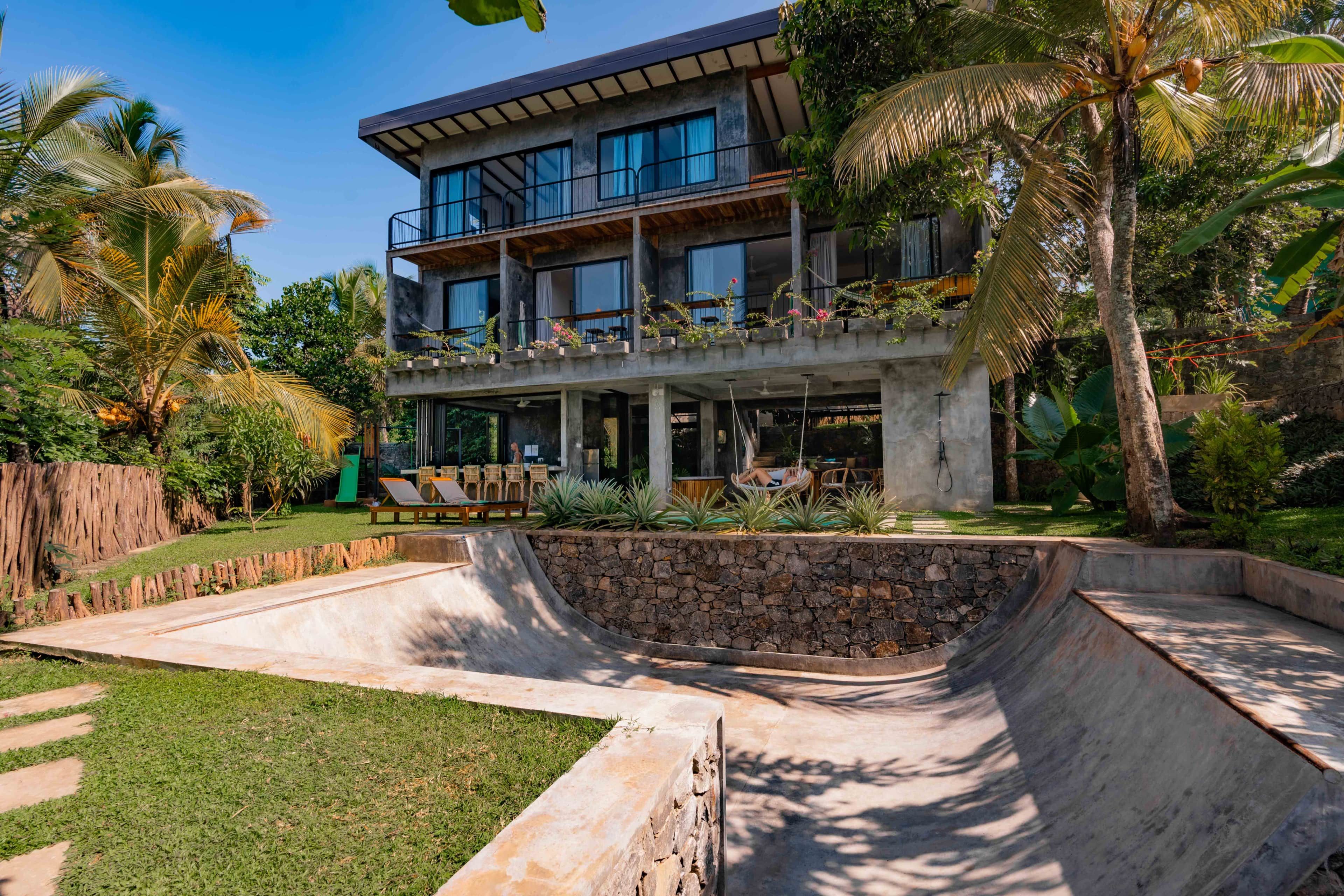 Um edifício moderno de dois andares com varanda, cercado por vegetação exuberante e uma rampa de skate de concreto.
