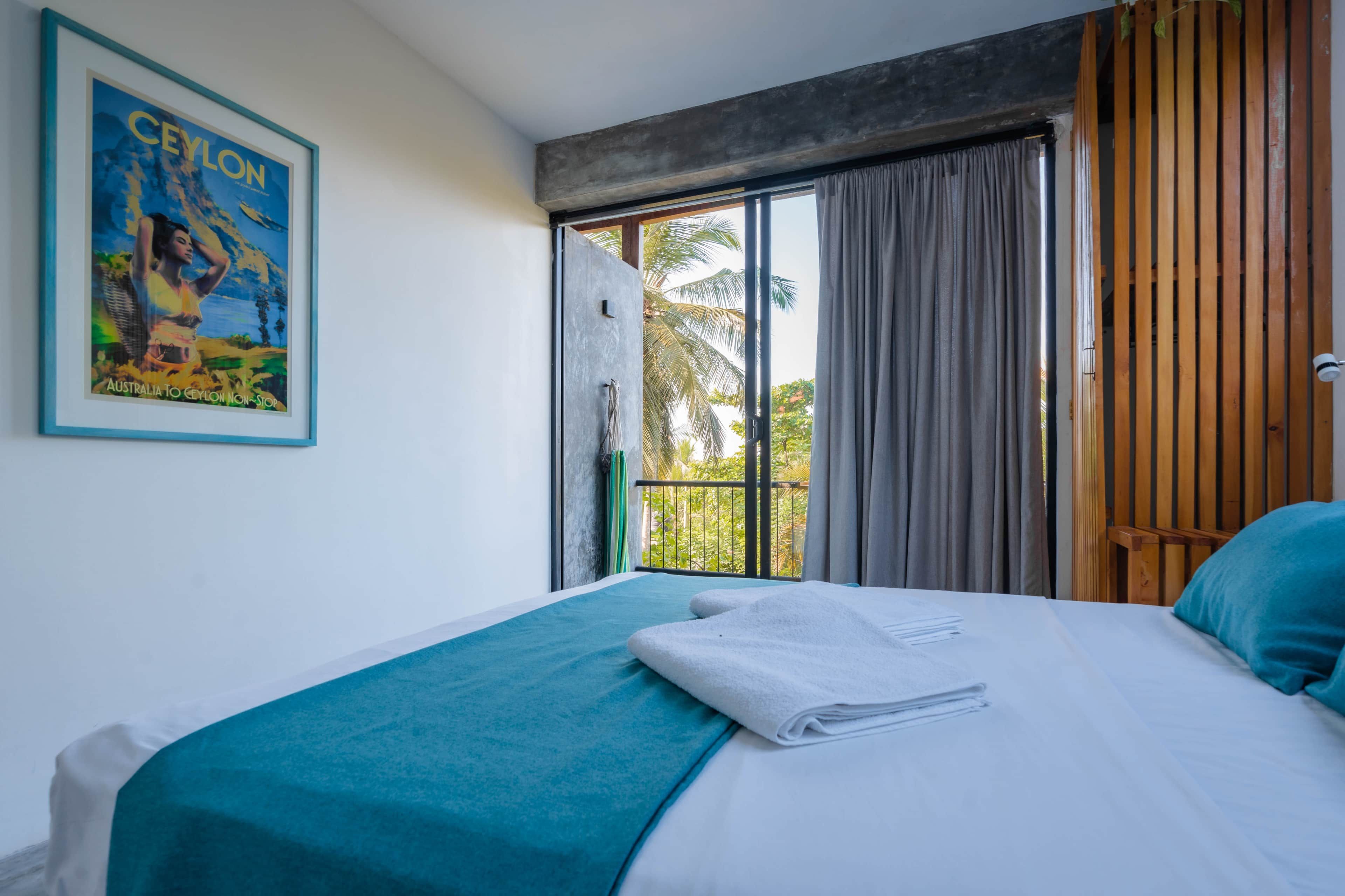 A hotel room with a bed, a window with curtains, a view of greenery outside, and a poster on the wall.