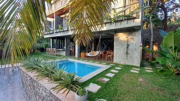 Area piscina esterna moderna con solarium in cemento, circondata da vegetazione lussureggiante e una sedia a dondolo.