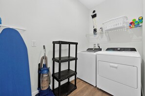 A laundry area with a washing machine, a dryer, a vacuum cleaner, and a black shelving unit. - GoUppers® Doppio | 5BR Townhome + Private Pool (Kissimmee)