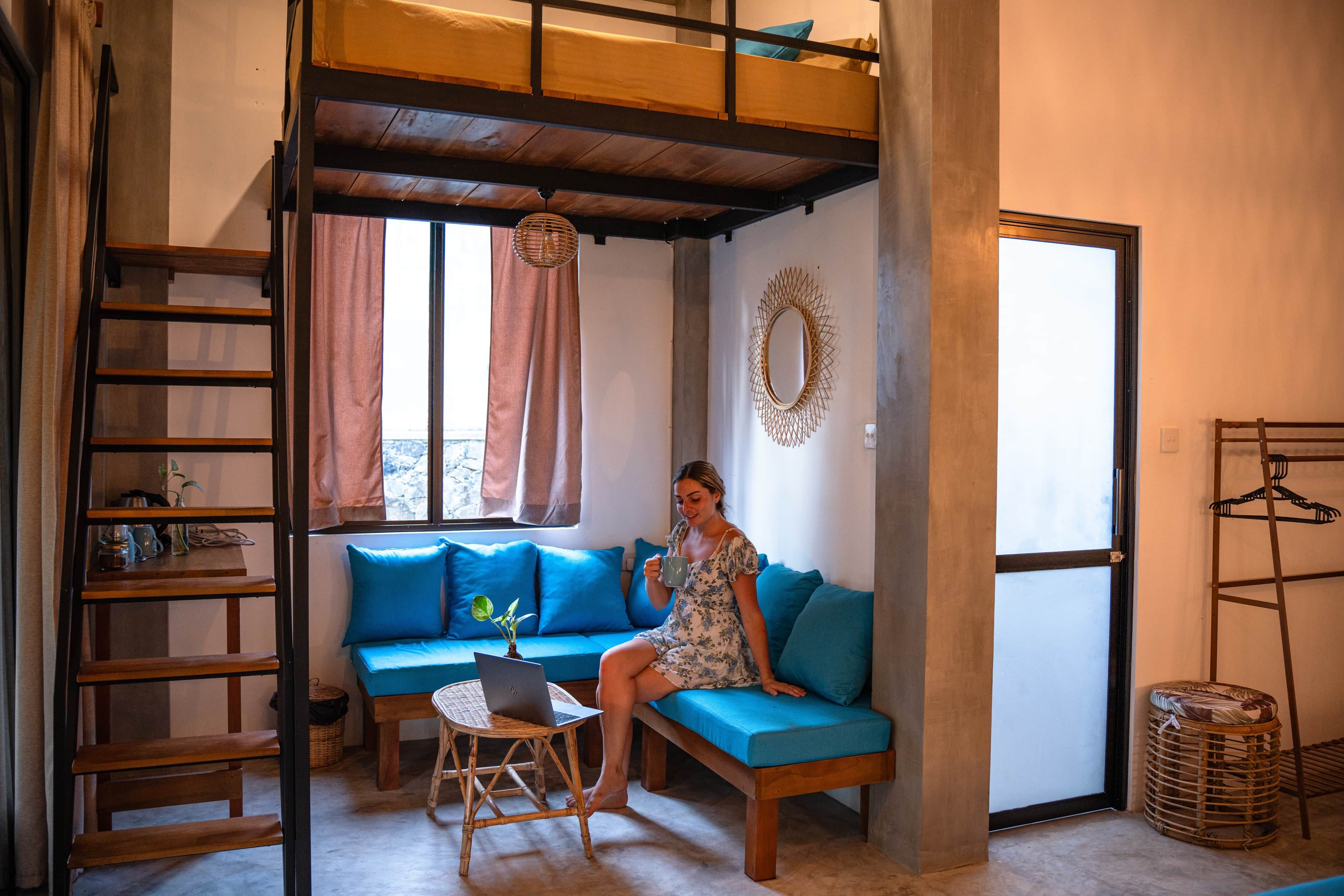 A room with a bunk bed, a sofa, a laptop on a stool, and a hanging light fixture.