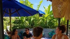 Eine Gruppe von Menschen sitzt unter blauen Sonnenschirmen an einem Holztisch, im Hintergrund üppiges Grün.