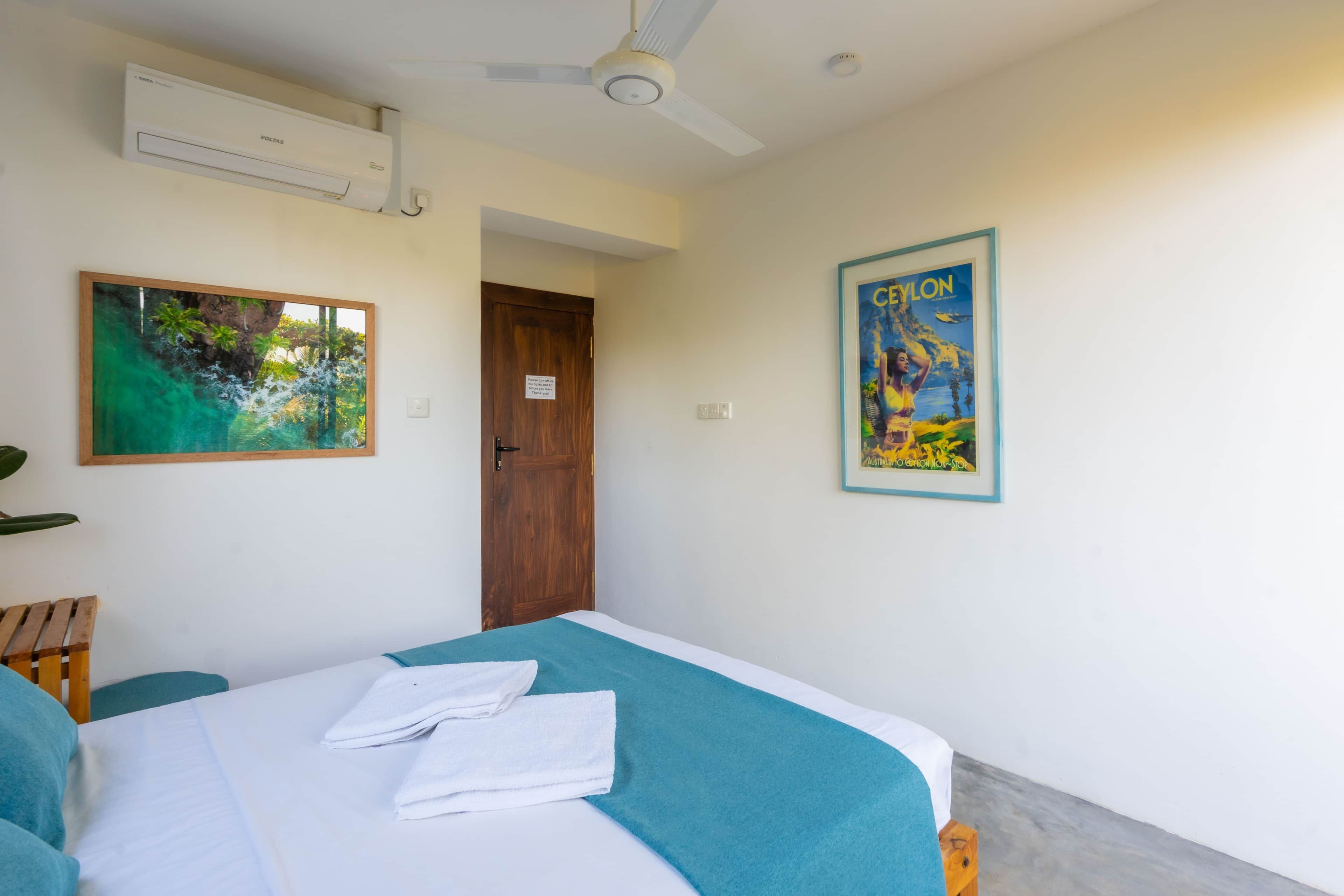 Une chambre d’hôtel avec un lit, un ventilateur de plafond, un appareil de climatisation, une porte et deux tableaux encadrés accrochés aux murs.