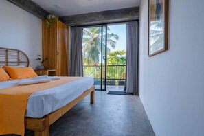Une chambre à coucher avec un lit, une tête de lit en bois, une table de chevet, une plante en pot et une vue sur des palmiers, visible à travers une porte-fenêtre coulissante.
