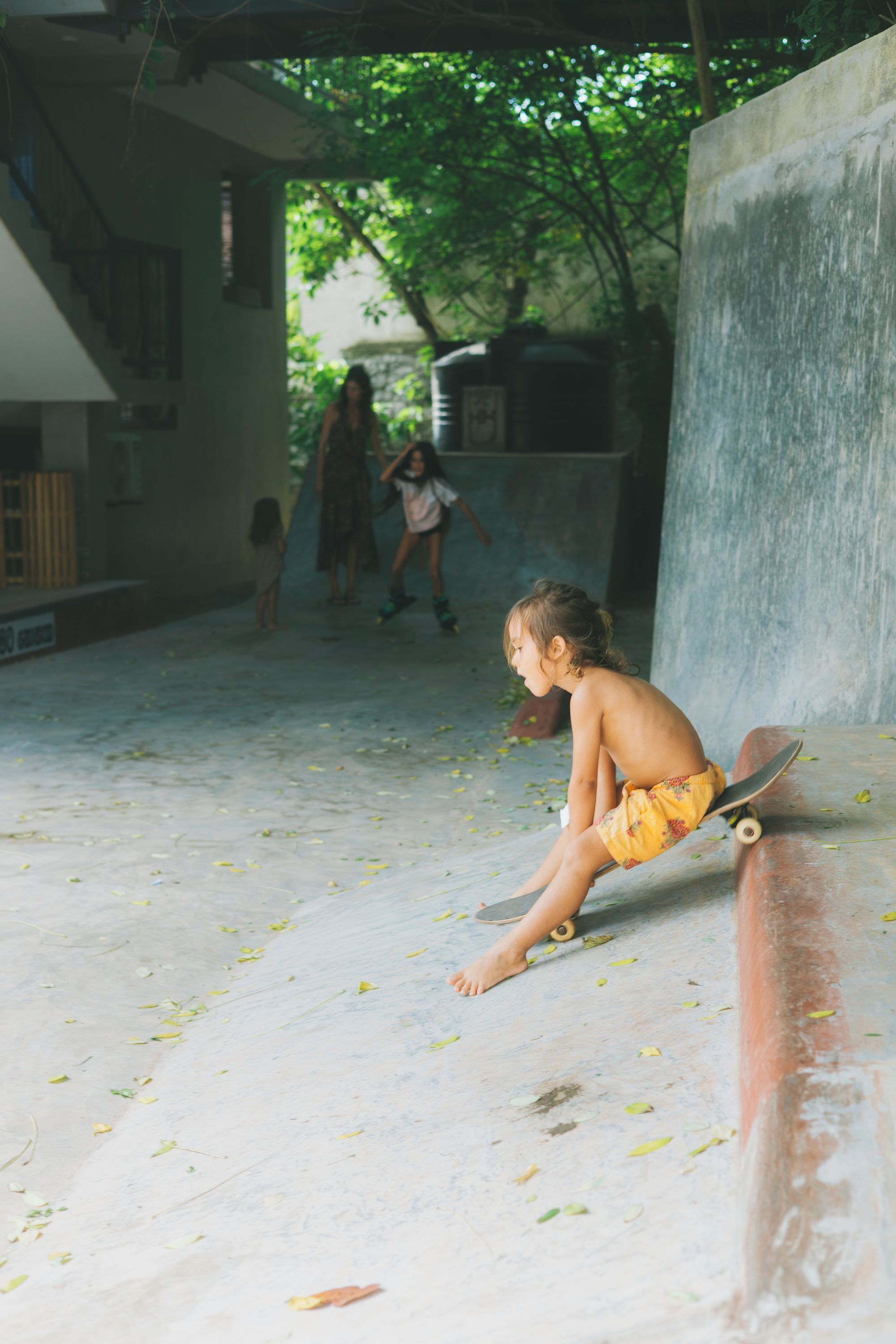 A child sitting on a skateboard in an outdoor area with a concrete floor, surrounded by greenery and a building structure.