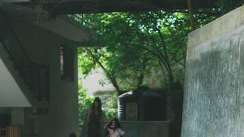 A child sitting on a skateboard in an outdoor area with a concrete floor, surrounded by greenery and a building structure.