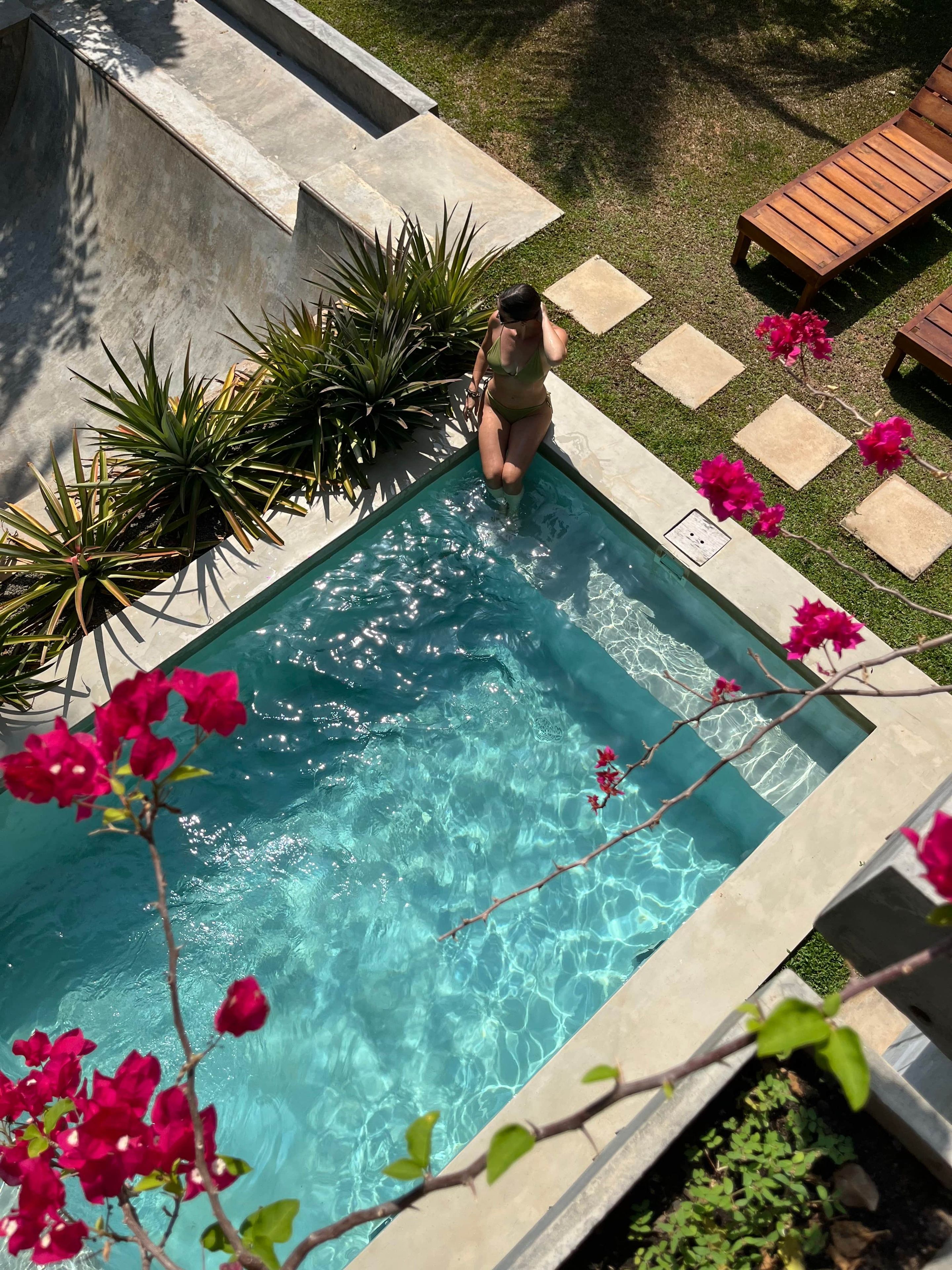 Una piscina con una persona in piedi sul bordo, circondata da piante e un pontile di legno con sdraio.