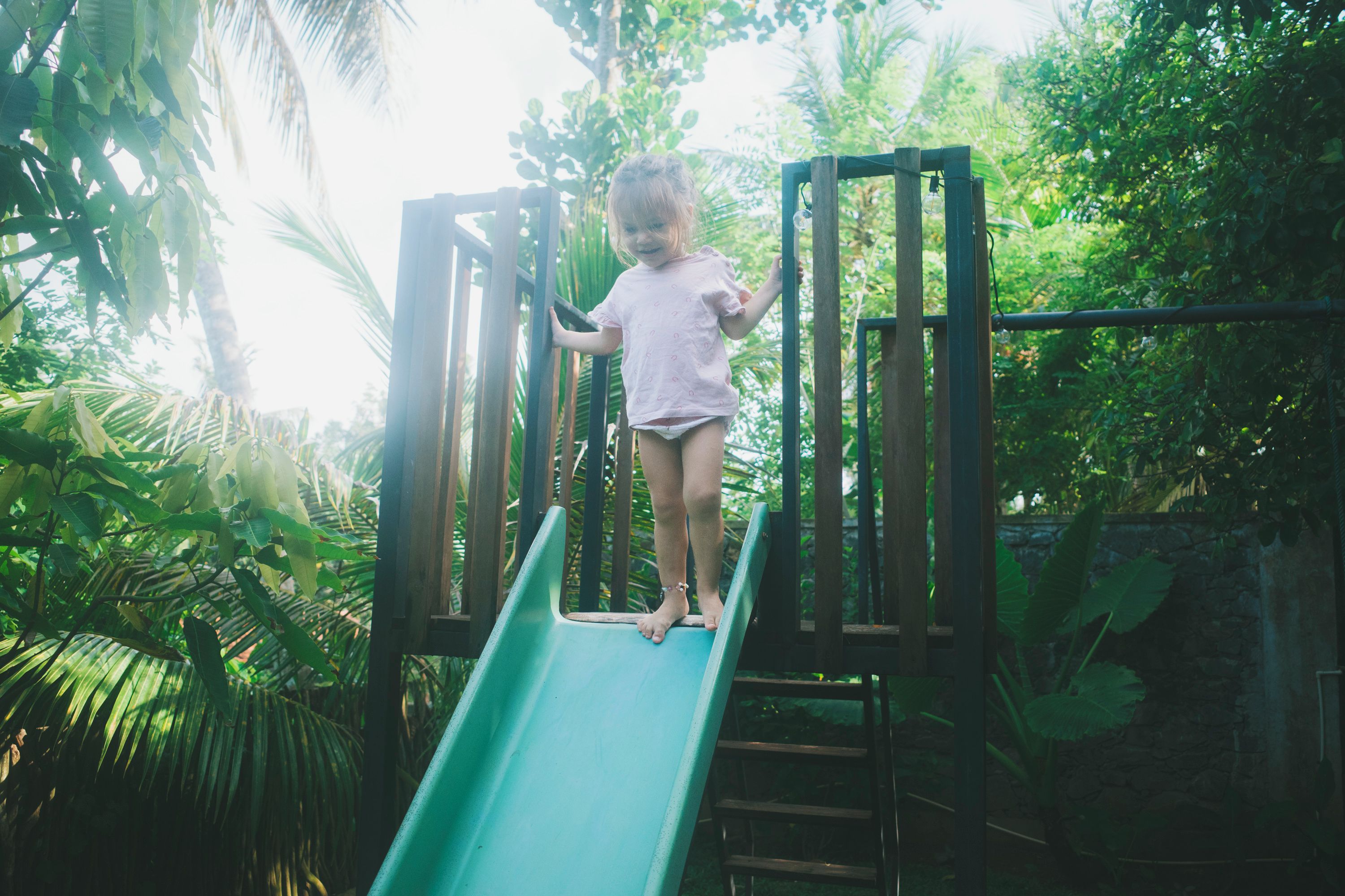 Un enfant sur un toboggan vert, dans un parc pour enfants entouré d’une végétation luxuriante.