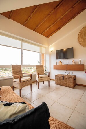 A living room with a high ceiling, a flat-screen TV mounted on the wall, wicker furniture, and a view through large windows.