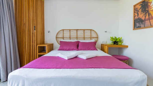 A bed with a white and pink bedspread, two pink pillows, and a white towel. There is a wooden headboard, a wooden nightstand with a plant, and a grey curtain. - The Orchid Room (Maaliyadda)
