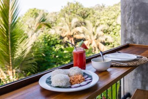 Uma bandeja com um prato de comida, um copo de bebida vermelha e uma tigela numa varanda com vista para o verde.