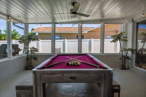 Une table de billard avec des boules et des queues, un ventilateur de plafond et des plantes en pot.