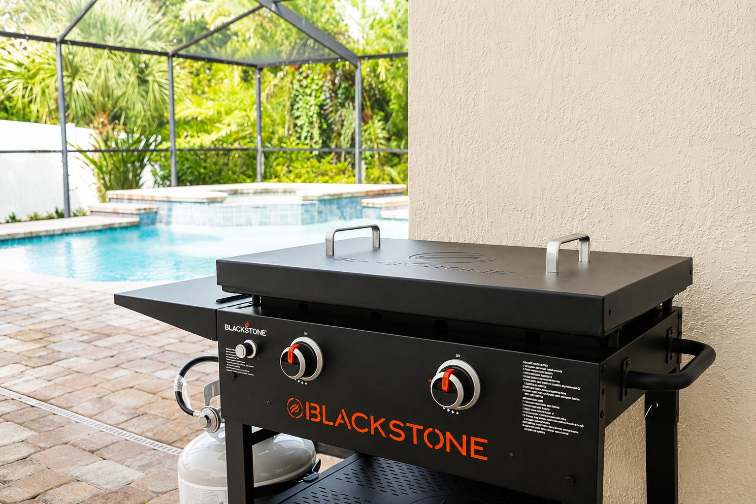 Una parrilla BLACKSTONE para exteriores con dos quemadores, un tanque de gas y una repisa lateral, cerca de una piscina.