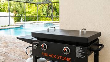 Una parrilla BLACKSTONE para exteriores con dos quemadores, un tanque de gas y una repisa lateral, cerca de una piscina.