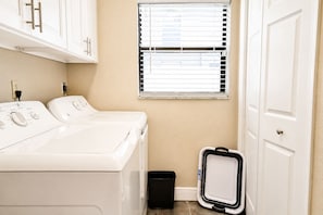 A laundry room with two white washers and dryers, a window with blinds, a black trash bin, and a white door.