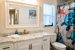 A bathroom with a marble countertop, a sink with a gold faucet, a white toilet, and a shower curtain with a marine design. - Serene Pool & Lake Retreat Sleeps8 Tarpon Springs (Tarpon Springs)