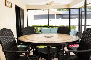 A round table with a mosaic top surrounded by wicker chairs, a ceiling fan, and a view of greenery outside.