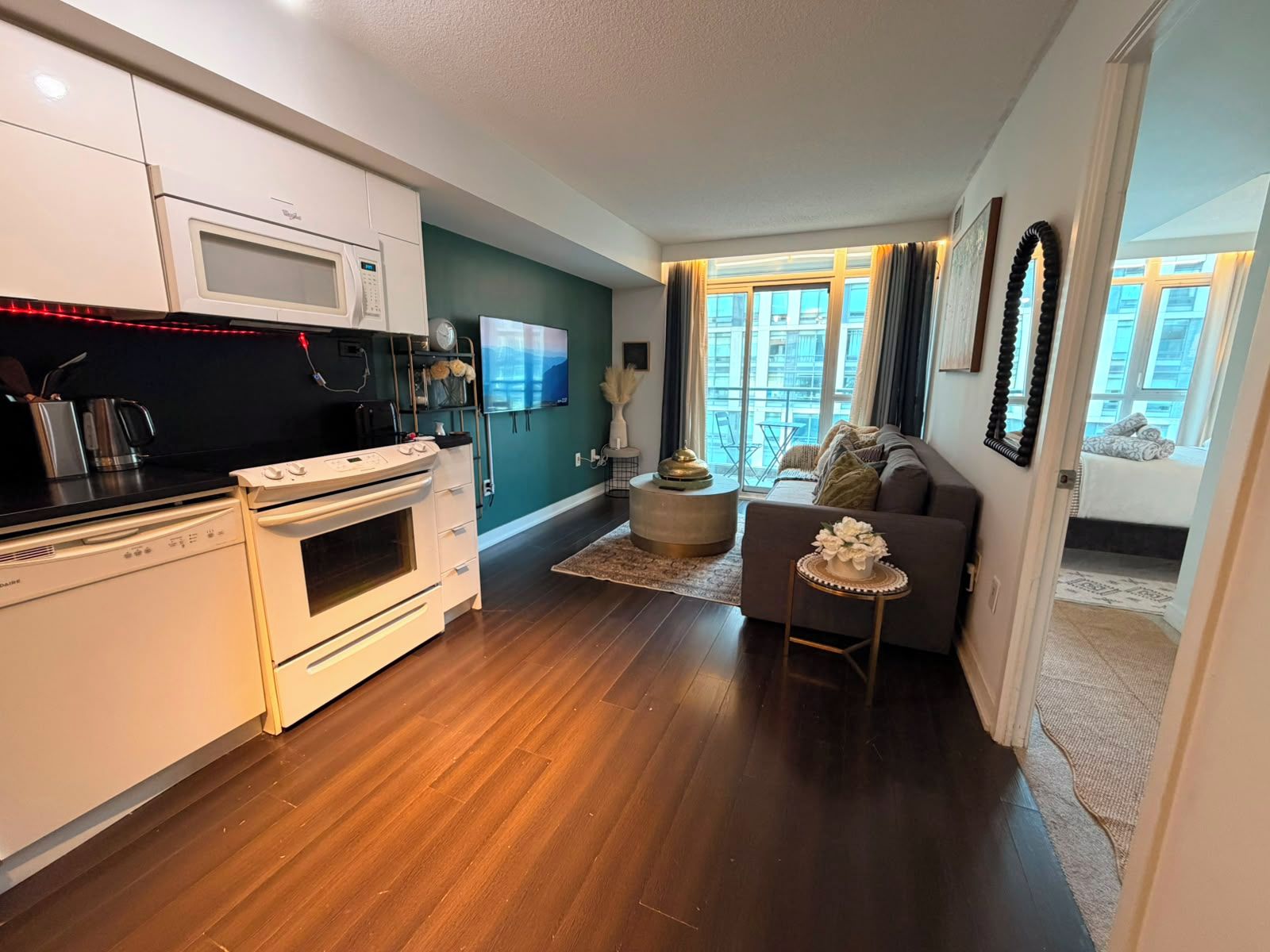 A modern kitchen with a microwave, stove, and coffee maker. There is a dining area with a sofa and a TV. The room has wooden flooring and large windows.