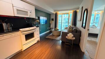 A modern kitchen with a microwave, stove, and coffee maker. There is a dining area with a sofa and a TV. The room has wooden flooring and large windows.