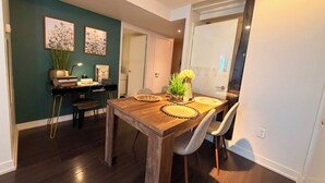 Un comedor con una mesa de madera, dos sillas y un jarrón de flores. Se observa una estantería con libros y una lámpara. Las paredes están pintadas en varios colores, y hay una puerta que da a otra habitación.