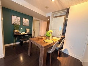 Un coin repas avec une table en bois, deux chaises et un vase de fleurs. On y trouve également une bibliothèque remplie de livres et une lampe. Les murs sont peints de couleurs différentes, et une porte donne accès à une autre pièce.