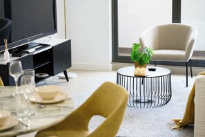 A modern living room with a flat-screen TV, a round coffee table with a plant, a white armchair, and a dining area with a table set for two. - Modern 1BR and Study Near Airport Shops & Parking (Botany)