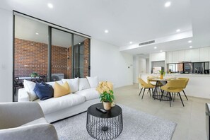 A modern living room with a sofa, coffee table, and a dining area with chairs and a table. - Modern 1BR and Study Near Airport Shops & Parking (Botany)