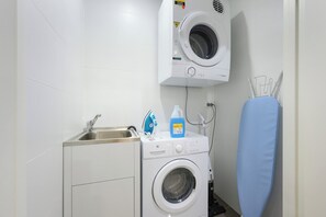 A compact laundry room with a washing machine, a sink, and a drying rack. - Modern 1BR and Study Near Airport Shops & Parking (Botany)
