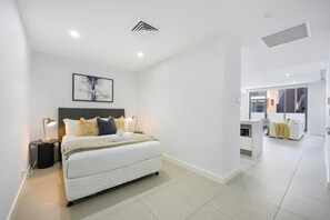 A modern bedroom with a large bed, bedside lamps, a painting, and a view of the living area. - Modern 1BR and Study Near Airport Shops & Parking (Botany)