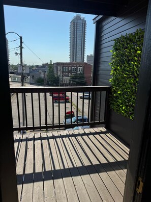 A wooden deck with a metal railing, a view of a parking lot, and a building with a green plant wall. - Historic Home Away From Home (Indianapolis)