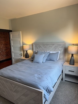 A bedroom with a bed, two bedside tables, a nightstand, a door, and a brick wall. - Historic Home Away From Home (Indianapolis)