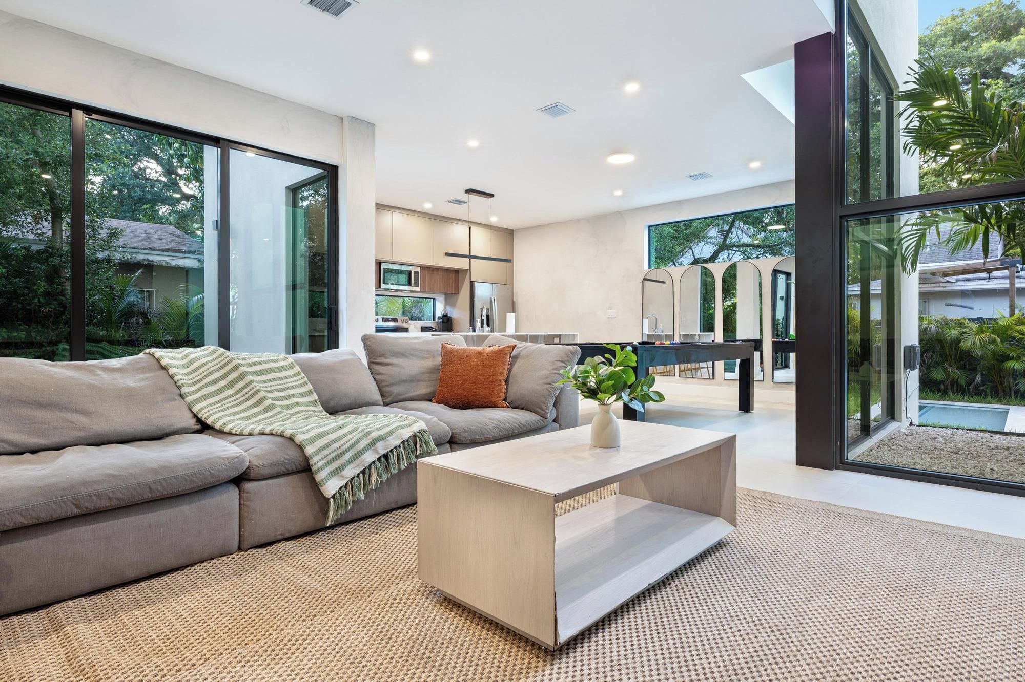 A modern living room with a sofa, coffee table, and a view of the kitchen and outdoor area.