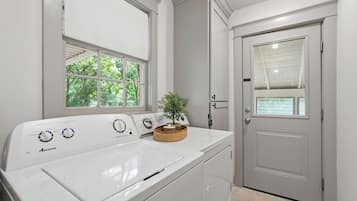 A laundry room with a front-loading washer and dryer, a window with a view of trees, and a door with a window.