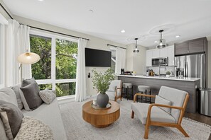 A modern living room with a sofa, armchair, and coffee table, a kitchen with stainless steel appliances, and large windows offering a view of greenery. - Stylish 3BR Seattle Retreat near UW w/ City Views (Seattle)