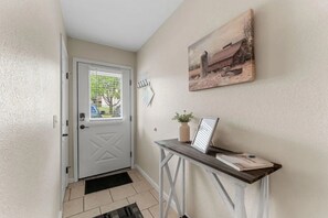 Een gang met een witte deur, een kleine tafel met een plant en een lijst met een foto aan de muur.