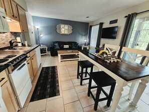 Eine moderne Küche mit einer zentralen Kochinsel, ein Essbereich mit einem dunklen Holztisch und ein Wohnzimmer mit einem schwarzen Sofa.