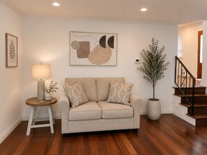 Ein Wohnzimmer mit einem beigefarbenen Sofa, einem Beistelltisch mit Lampe, einem großen abstrakten Gemälde, einer Topfpflanze und einer Treppe.
