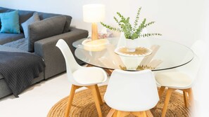 Ein modernes Wohnzimmer mit Glastisch, weißen Stühlen, einem grauen Sofa und einer Topfpflanze.