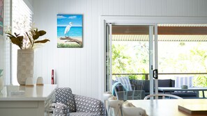 A room with a white wall, a painting of a bird on a branch, a white vase with flowers, and a sliding glass door leading to an outdoor area with a view of greenery. - Tropical Retreat on Reid (Wongaling Beach)