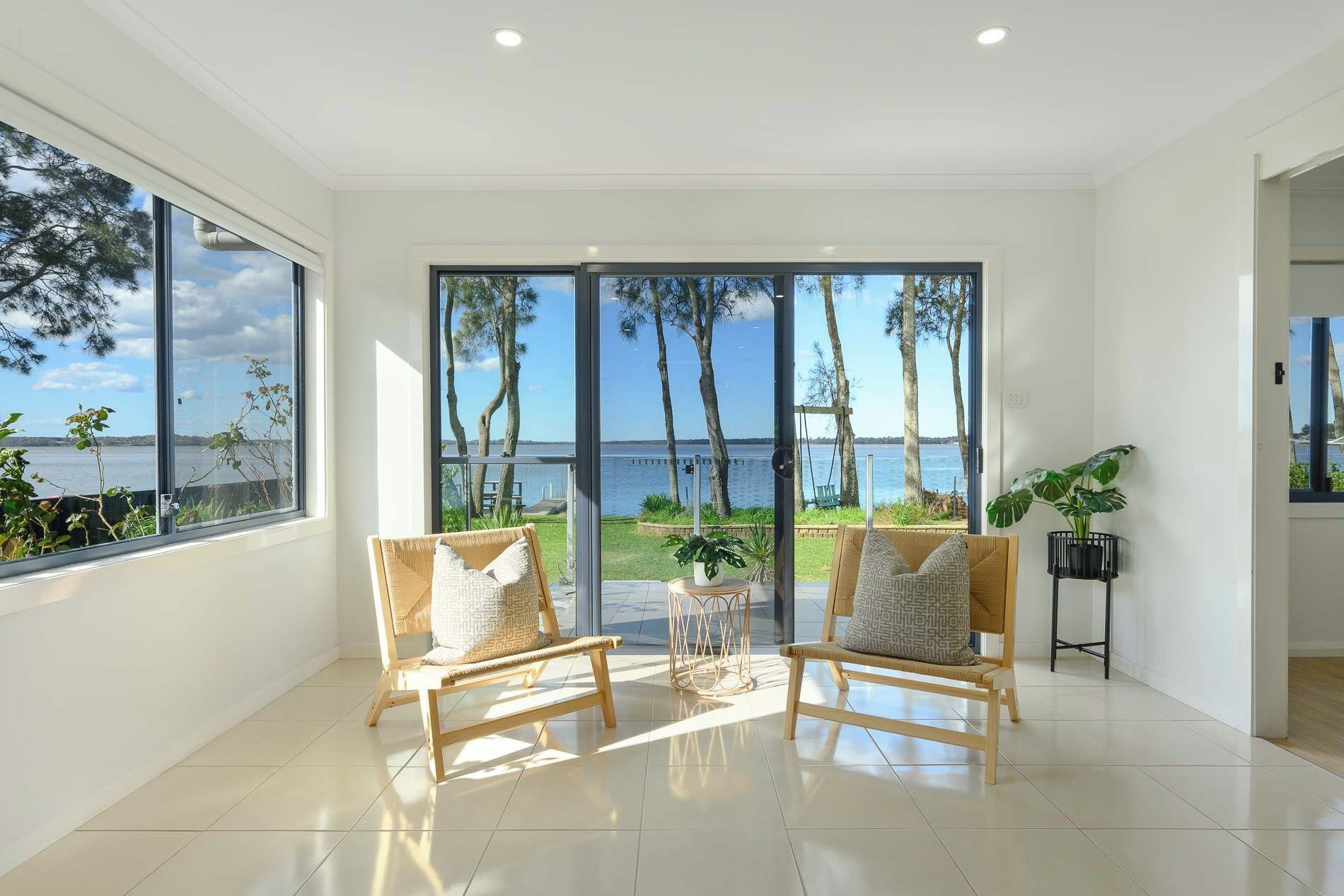 Amplia habitación con ventanas grandes que ofrecen vista al mar y palmeras. Hay dos sillas de mimbre con cojines y una pequeña mesa con una planta.