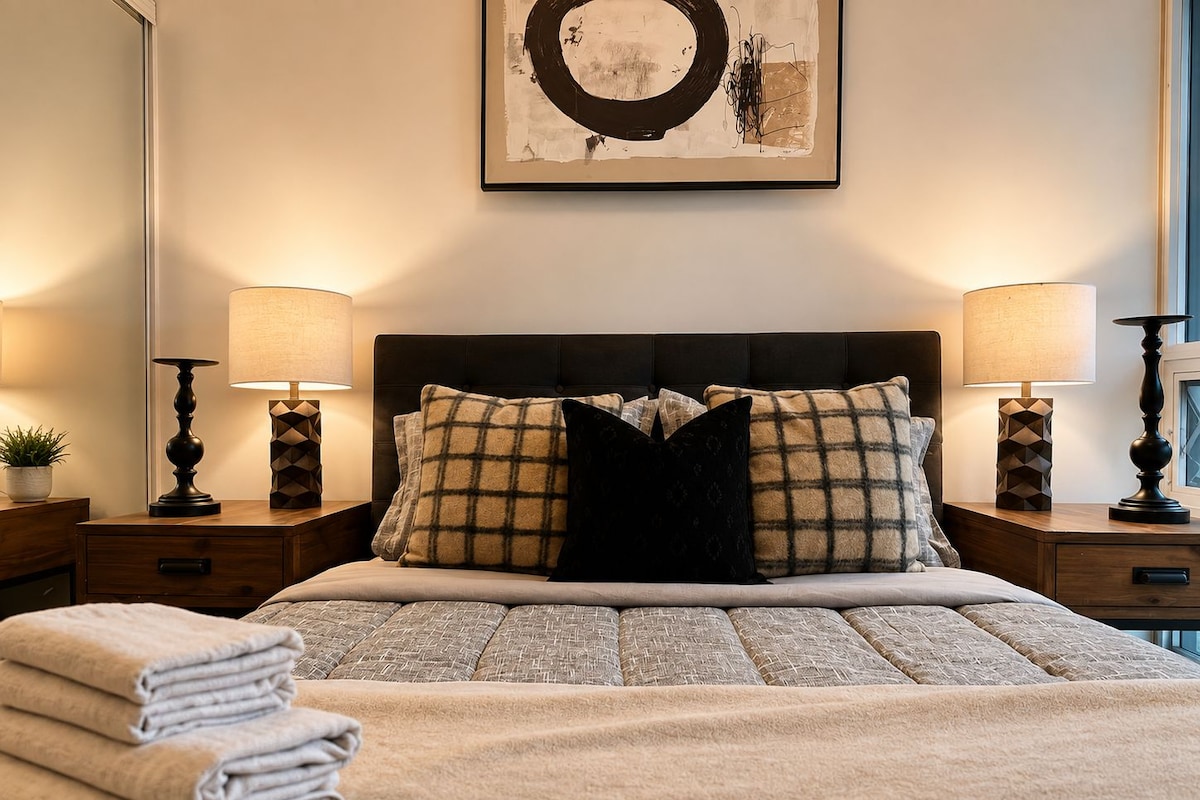 Un lit bien fait, avec une tête de lit, deux tables de chevet avec des lampes et une œuvre d’art accrochée au mur.