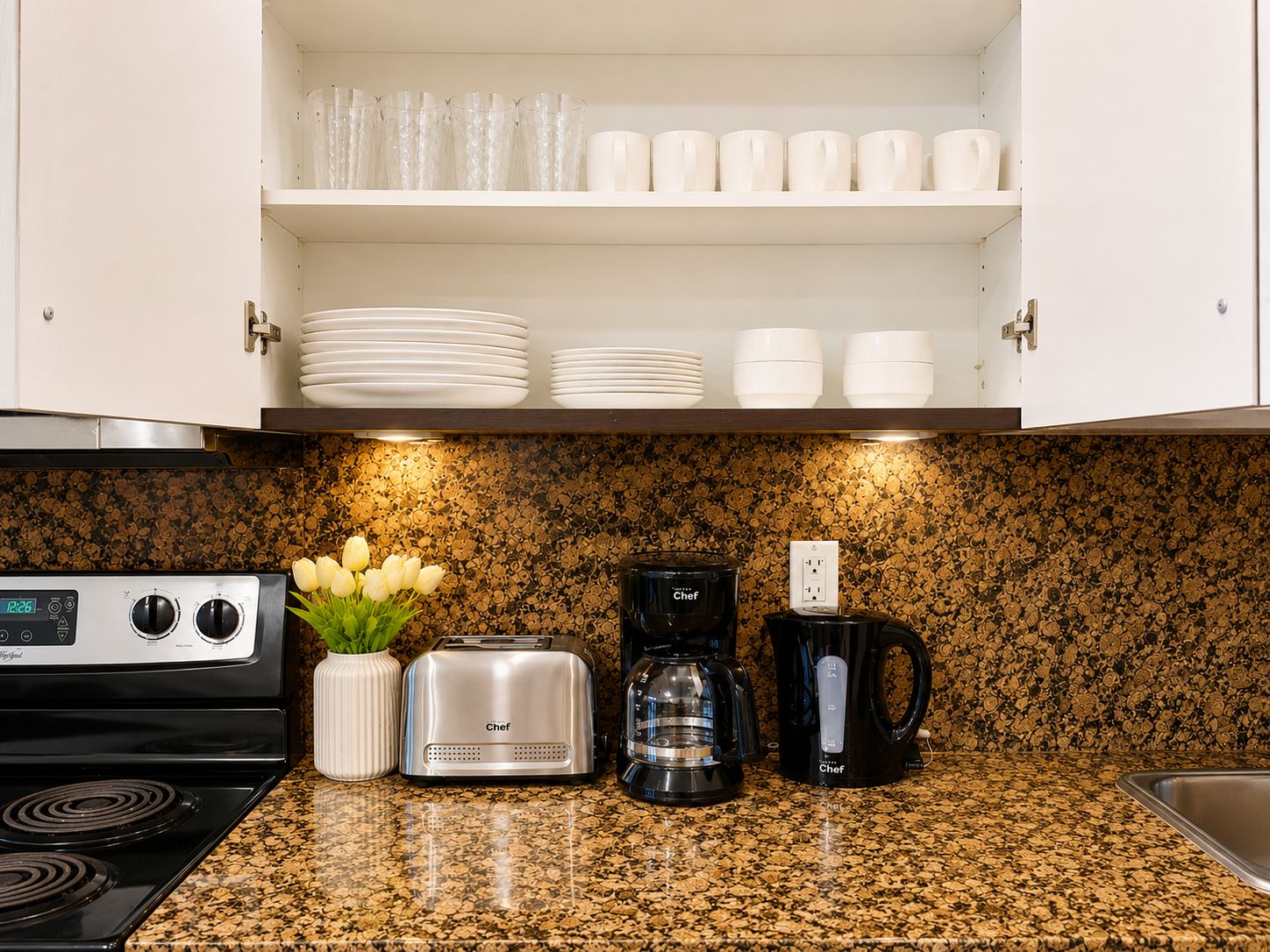 A kitchen with a stove, toaster, coffee maker, and a shelf with plates and cups.