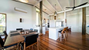 A modern kitchen with white cabinetry, a central island, and a dining area with a wooden table and chairs. - Island Vista Retreat (South Mission Beach)