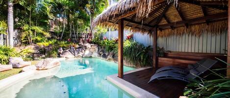 A pool area with a thatched-roof structure, lounge chairs, and tropical plants.