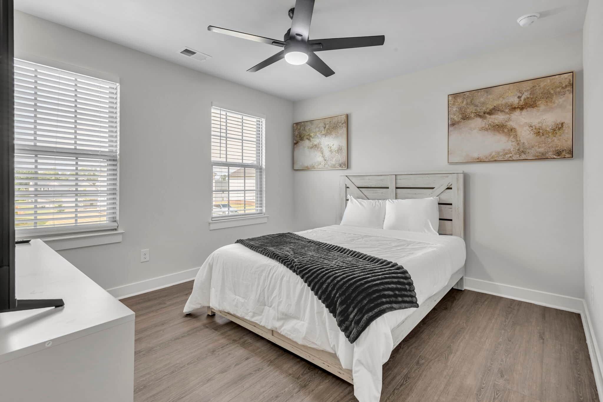 Quarto com cama, ventilador de teto, duas janelas com persianas e duas obras de arte na parede.