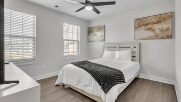 Quarto com cama, ventilador de teto, duas janelas com persianas e duas obras de arte na parede.