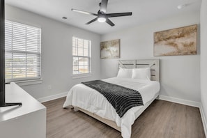 A bedroom with a bed, a ceiling fan, two windows with blinds, and two wall-mounted artworks.