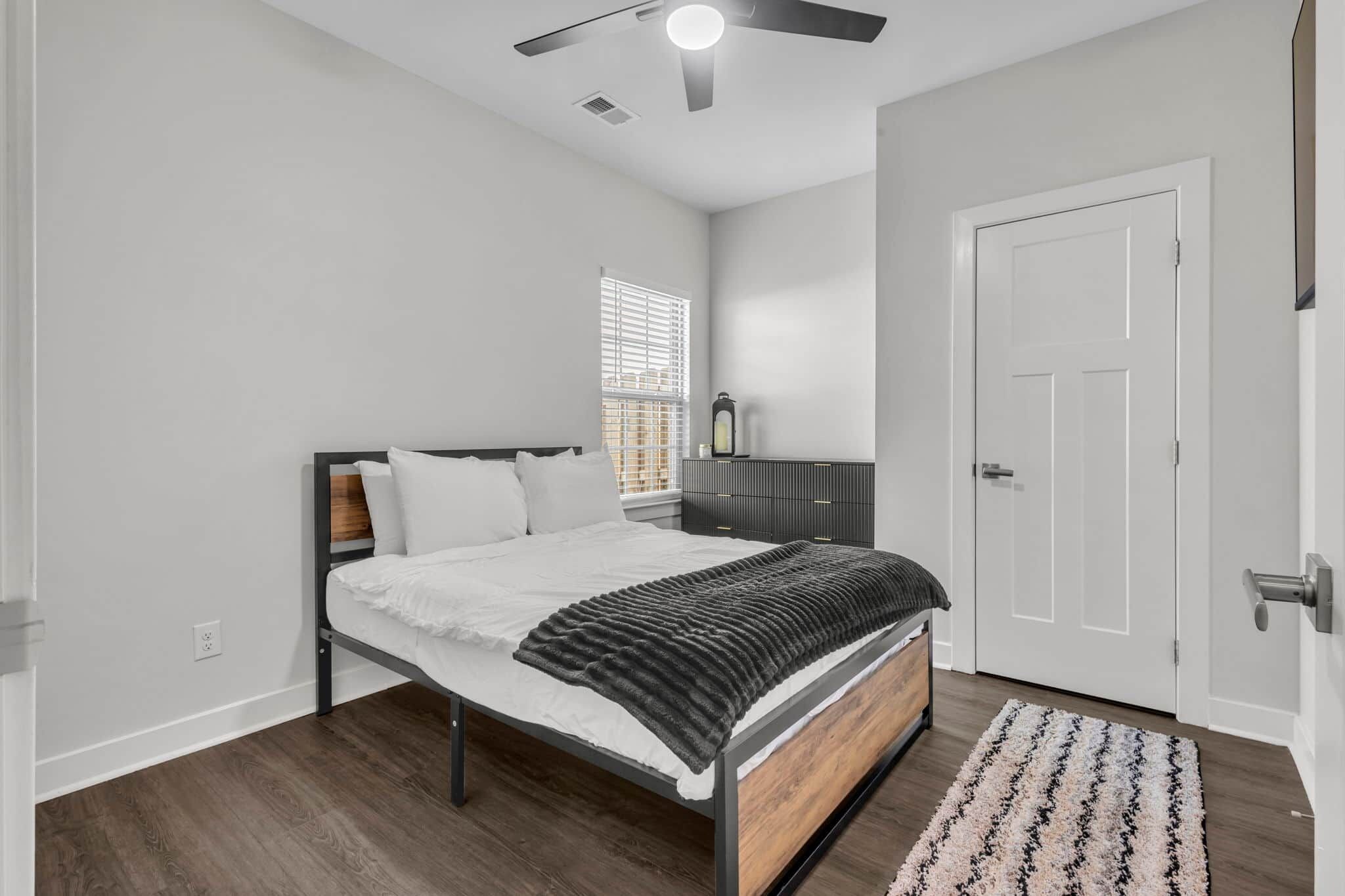 Une chambre à coucher comprenant un lit, une table de chevet, une fenêtre avec des stores, une porte et un ventilateur de plafond.