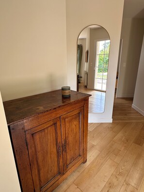 A wooden cabinet with a candle on top, a hallway with wooden flooring, and a door leading to a bright room with a view of greenery. - Country house with a pool between Bordeaux and St-Émilion (Croignon)